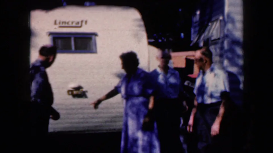 A woman in a blue patterned dress gestures while adults stand near the white Lincraft trailer, captured in soft 8mm color.