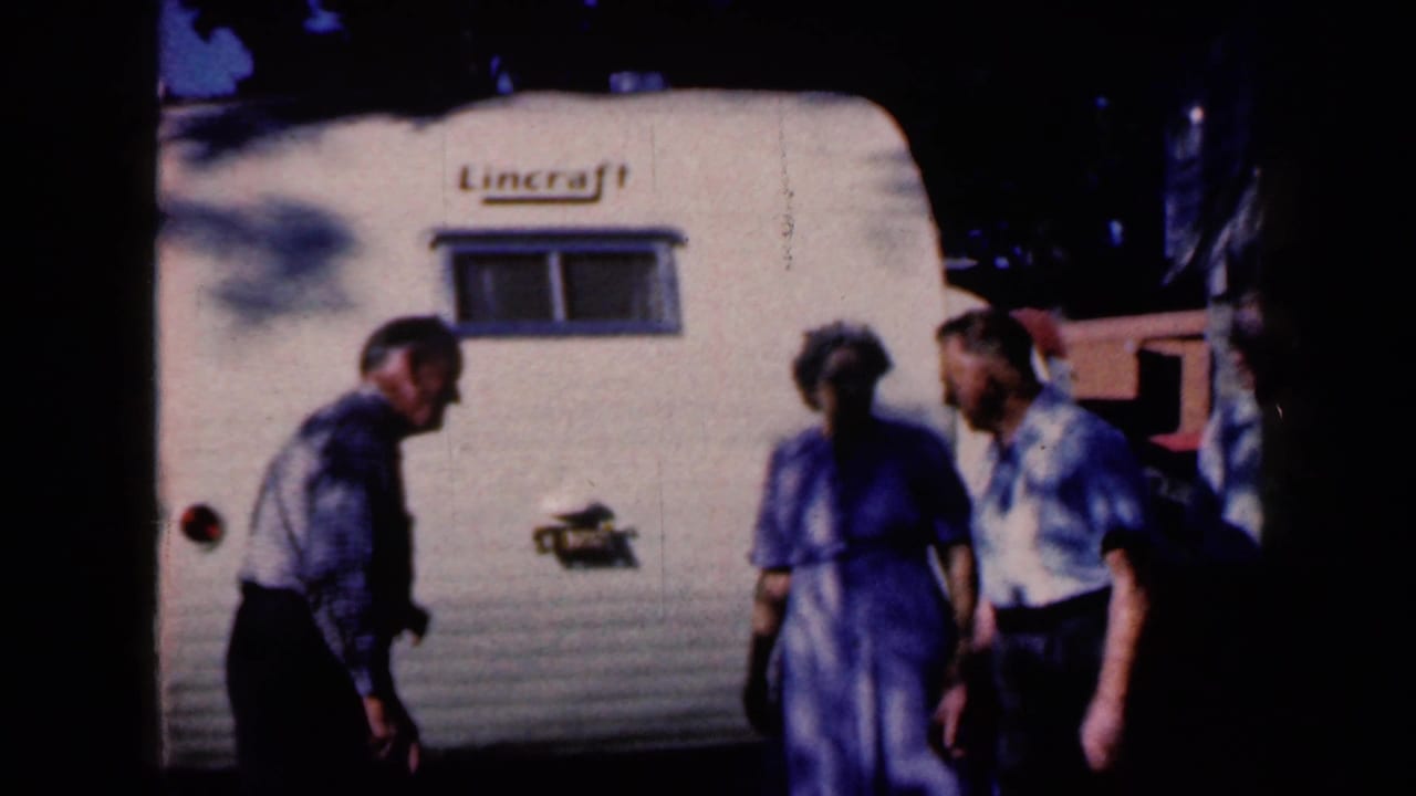 Adults stand beside a white Lincraft travel trailer in shaded outdoor light.