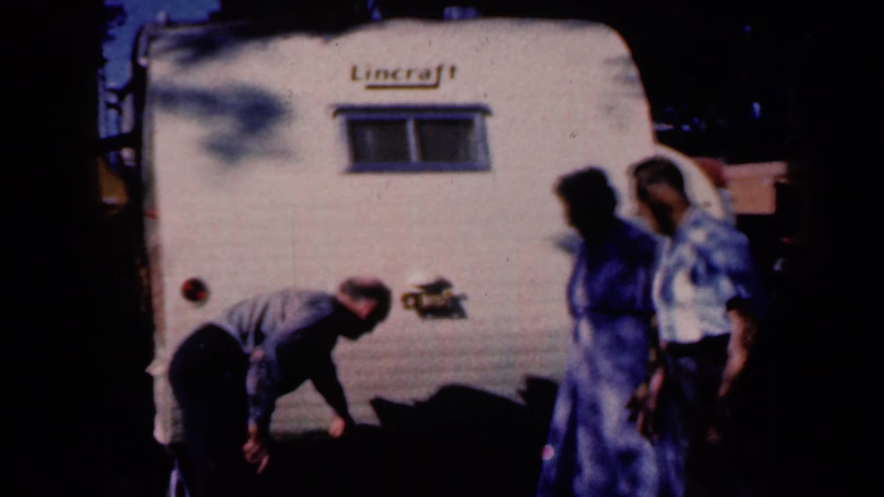 A man appears to inspect the wheel area of the white Lincraft trailer while others stand nearby in the Iowa 1959 family scene.