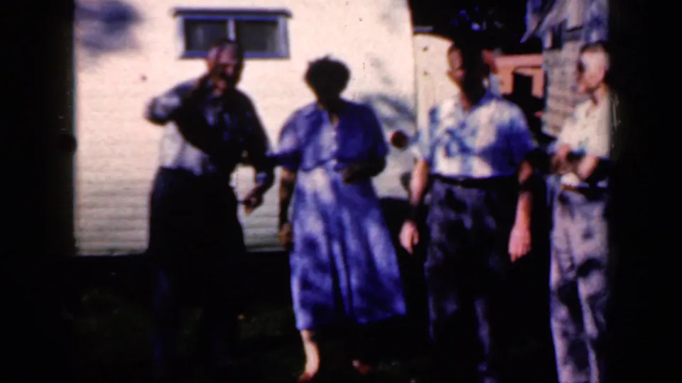 Four adults gather near the Lincraft trailer in the Iowa 1959 home-movie scene, with one man gesturing near his face in deep shade.