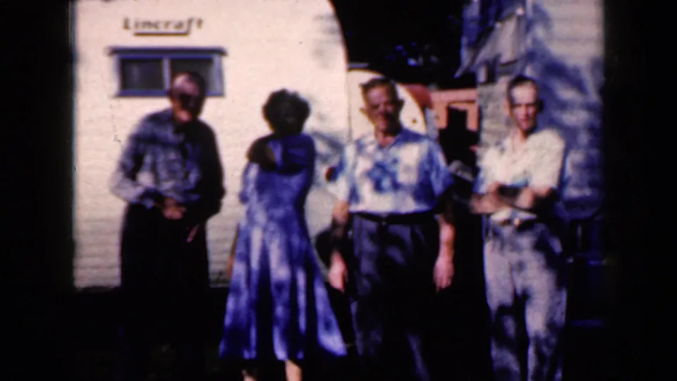 The group remains outside the white trailer in Iowa, with the woman in a blue dress turned toward the men.