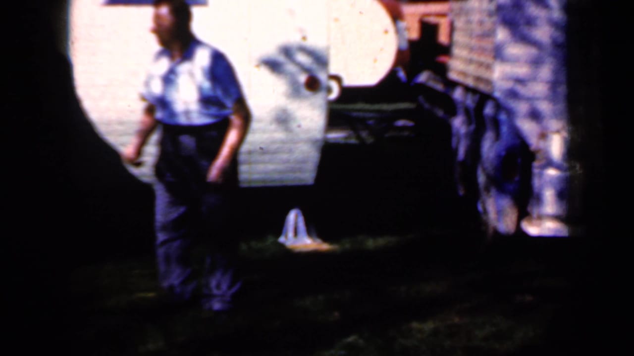 Man in a patterned shirt walks beside a white trailer in a shaded grassy yard.