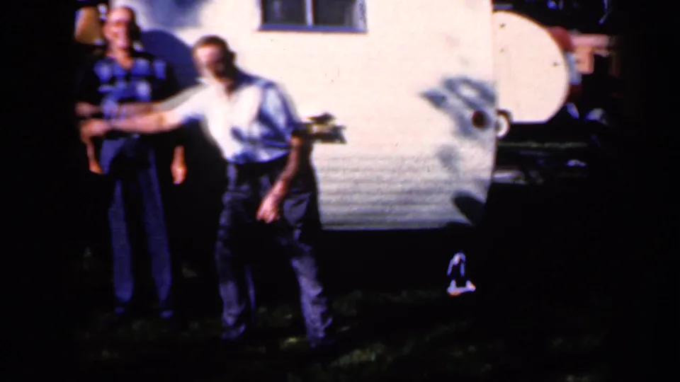 Two adult men stand outside a white trailer in Iowa, with one extending an arm as the greeting begins.