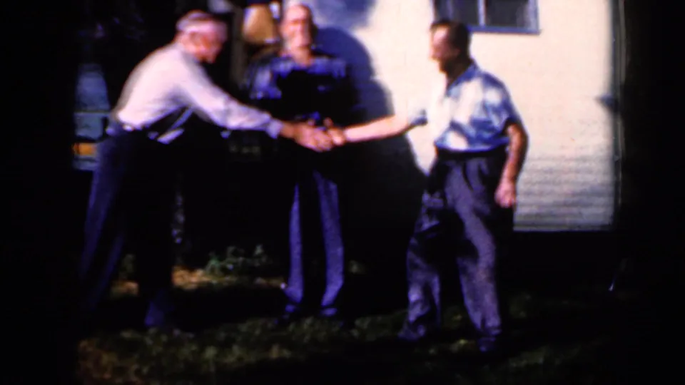 Three adult men gather in the yard near the trailer, with two men shaking hands in the center of the frame.