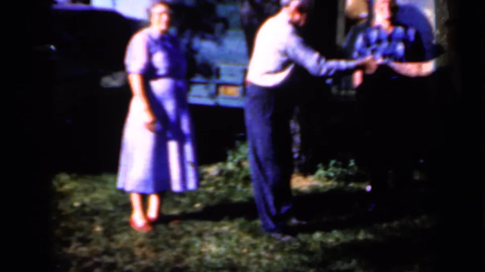 A woman in a purple dress stands nearby while two men complete a handshake in the shaded Iowa yard.