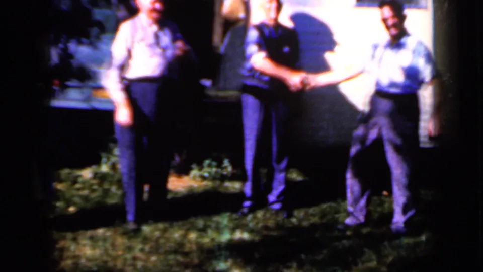 1959 Iowa home-movie frame of men standing at night on grass beside a pale exterior wall under strong light.