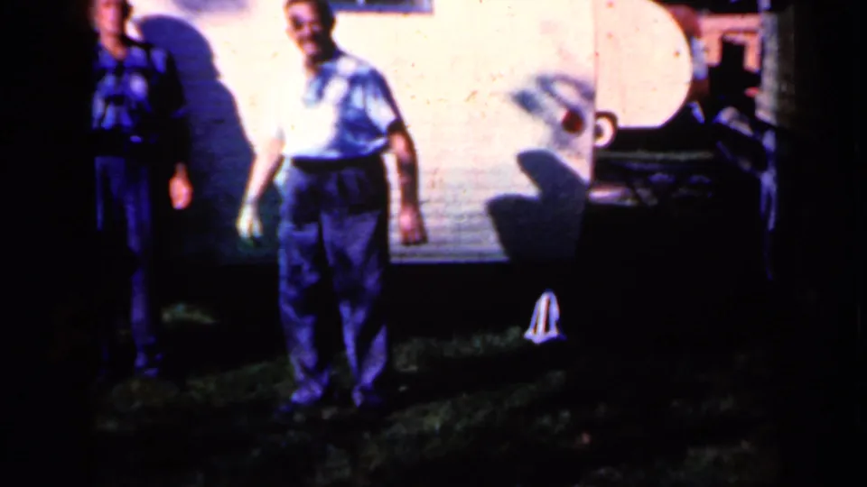 1959 Iowa home-movie frame showing a man facing the camera outdoors at night, with other adults partly visible near a wall.