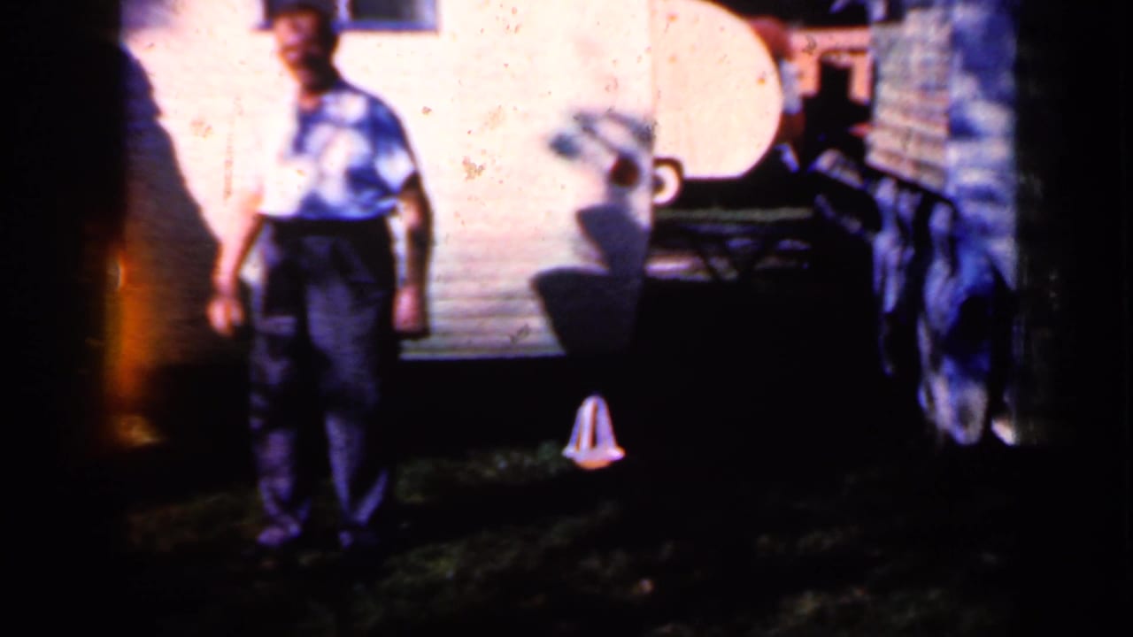 In Iowa 1959 home-movie footage, a man in a patterned shirt stands outside near a light trailer with yard shadows around him.