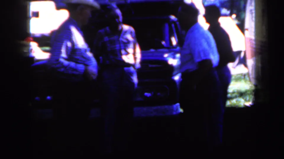 A close home-movie view shows men standing near the front of a dark truck during an outdoor Iowa gathering in 1959.
