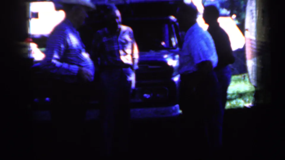 Men continue talking near a dark vintage truck, with bright grass and a building edge visible in the 1959 Iowa home-movie frame.