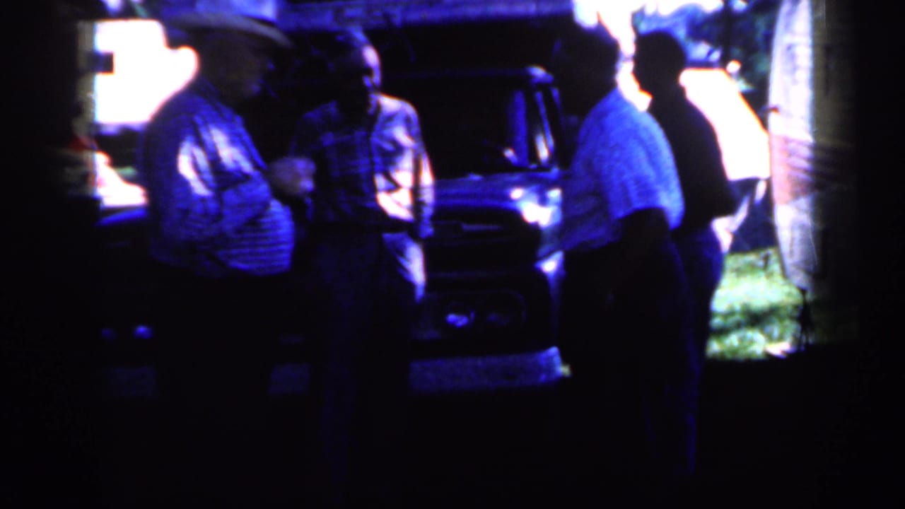 Adult men standing in shade near a dark truck and trailer outdoors.