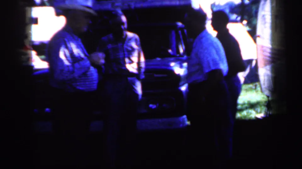 In Iowa home-movie footage from 1959, several adult men stand talking near a dark truck with grass and a trailer behind them.