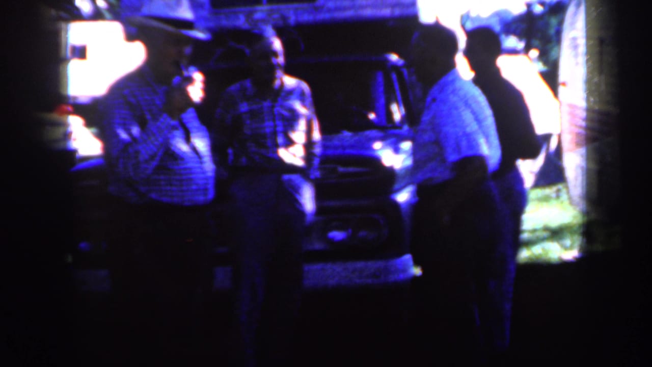 Several adult men appear to converse beside a parked vehicle in rural Iowa, with bright sun behind them and a trailer edge nearby.