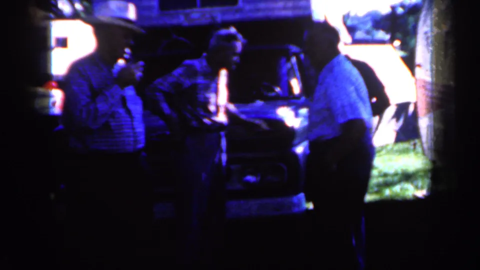 Adult men in mid-century casual work shirts stand near the front of a dark truck, filmed with strong blue cast and dense shadow.