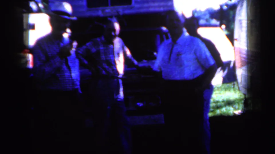 A man in a light shirt faces other men beside a truck in 1959 Iowa home-movie footage, with grass and a trailer in the background.