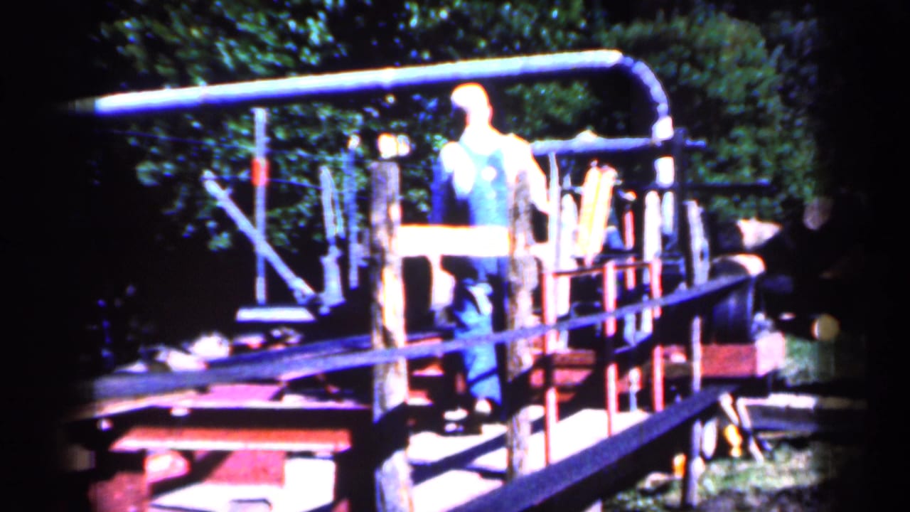 A man in overalls stands on a wooden and metal outdoor contraption.