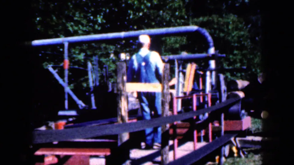 The man in overalls faces away while standing among rails, boards, and metal parts in a wooded backyard setting in Iowa home-movie footage.