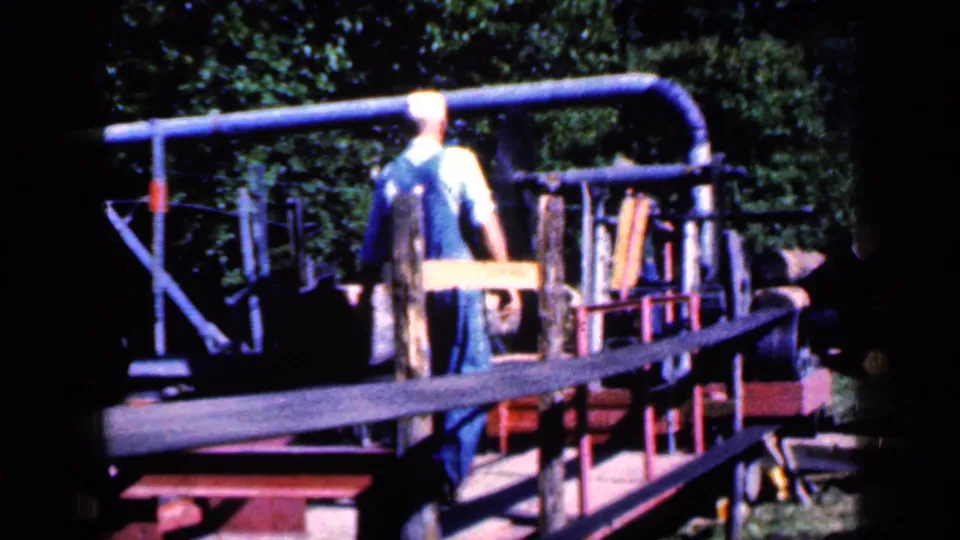 A man in overalls stands beside a rail-like wooden and metal structure in a wooded Iowa yard, with strong sun and 8mm color shift.