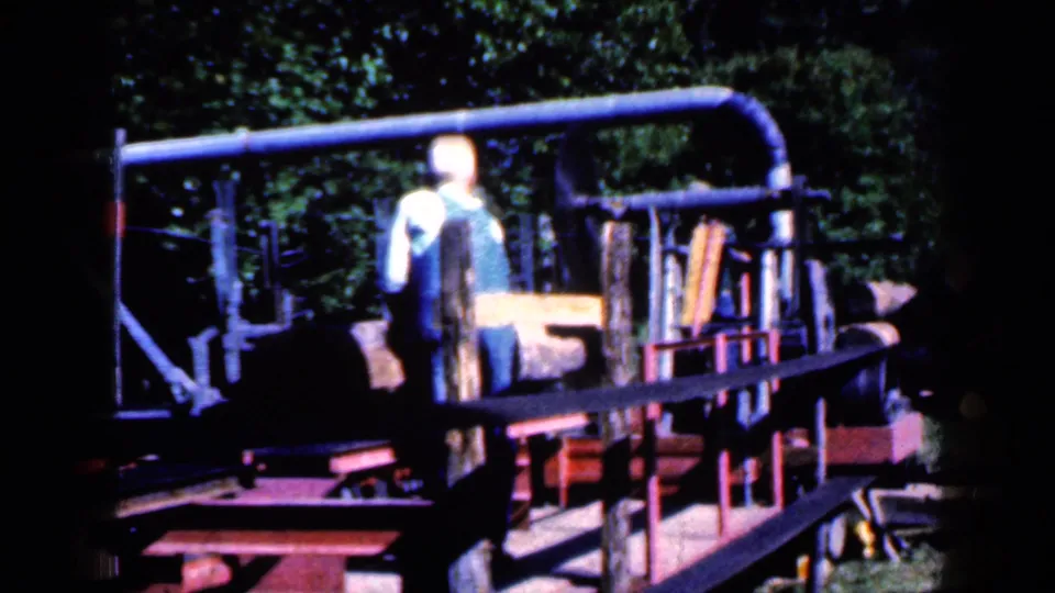 In 1959 Iowa footage, a man in a white cap and blue overalls stands near belt-driven lumber equipment outdoors.