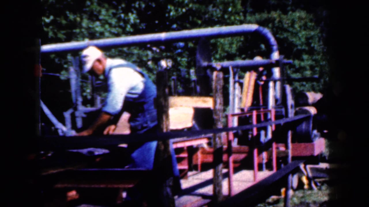 Man bending over an outdoor lumber machine with boards and rails nearby.
