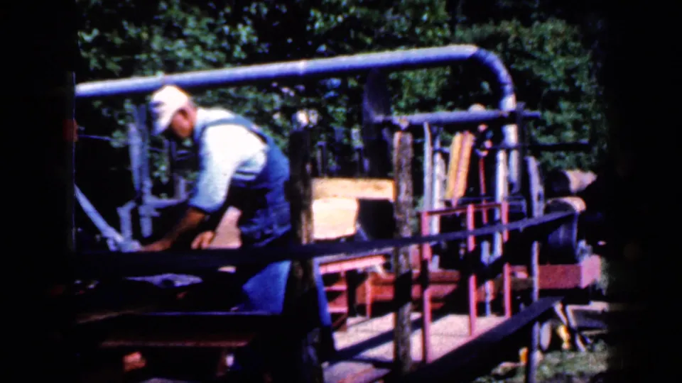 The worker bends toward the belt-driven lumber machine in a rural outdoor Iowa setting, with boards and trees visible.