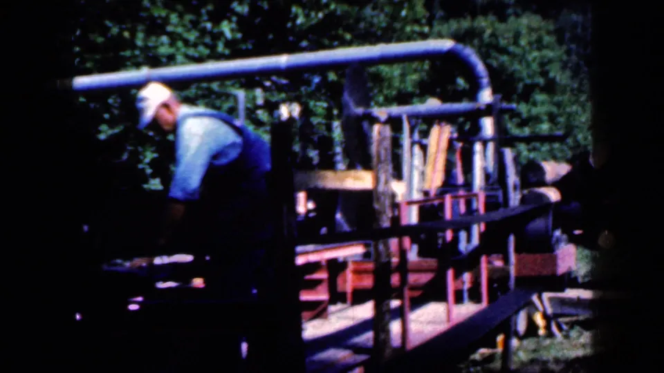 A man in work clothes moves beside the lumber machine, partly obscured by shadow and the contact-sheet frame edge.