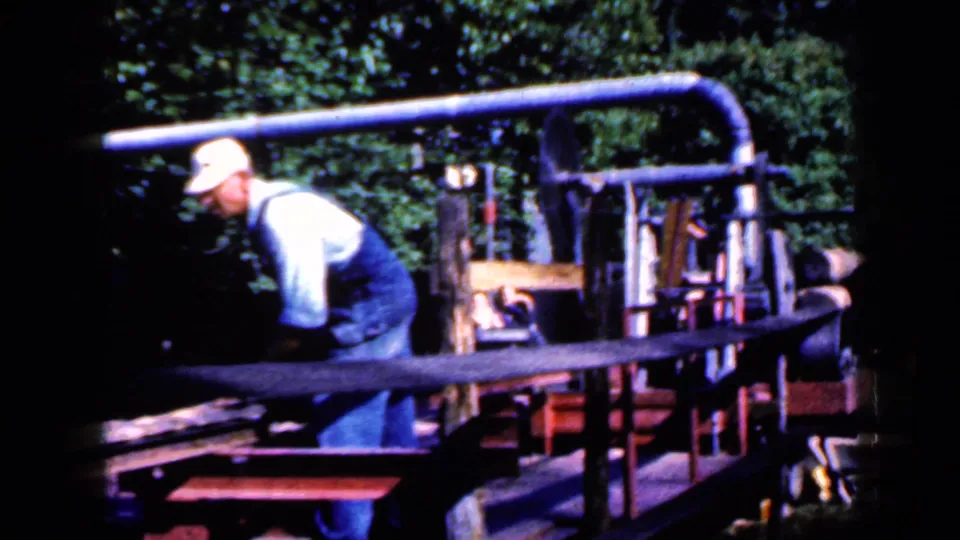 The 1959 Iowa home-movie frame shows the worker beside the red-framed machine with boards, rails, and trees in view.