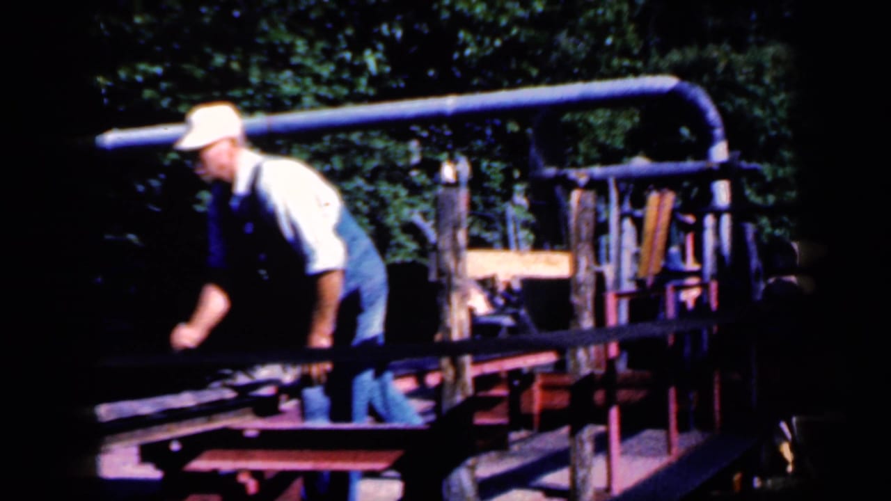 Man in overalls and a light cap leaning over outdoor machinery near wooden rails.