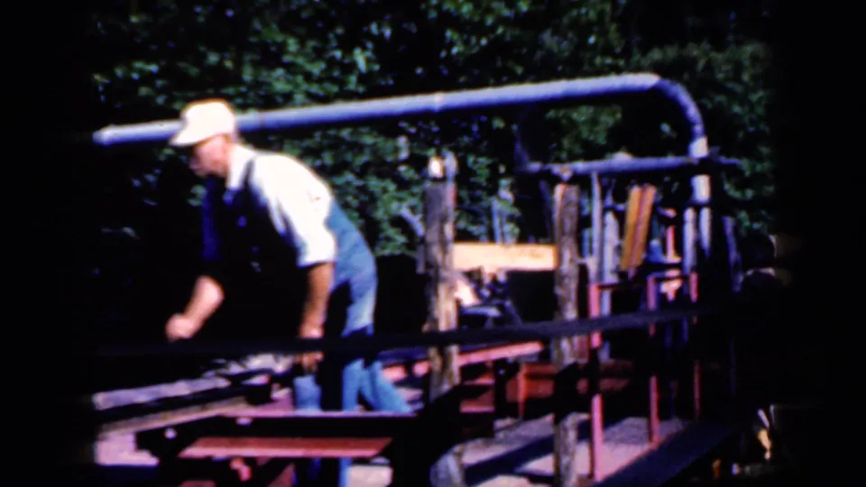 In Iowa 1959 home-movie footage, a man in overalls and a light cap tends outdoor machinery beside rails and trees.