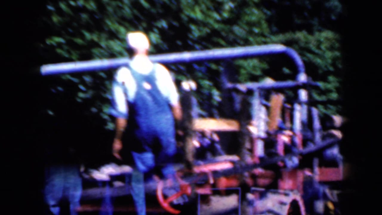 A man in blue overalls and a pale cap walks beside heavy outdoor machinery in the Iowa 1959 clip.