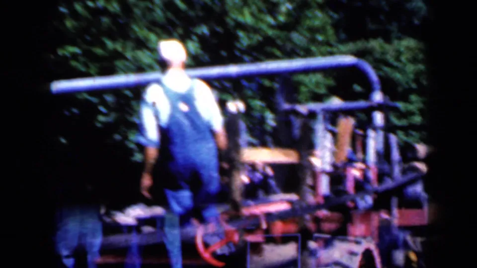 A man in blue overalls and a pale cap walks beside heavy outdoor machinery in the Iowa 1959 clip.