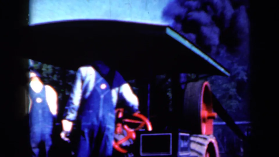 Two overall-clad men stand by a dark steam-powered machine with a red handwheel in Iowa, 1959.