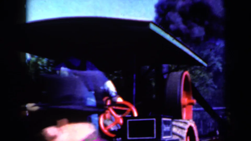 A man in overalls stands partly under the machinery housing near a red handwheel as smoke or steam darkens the frame.
