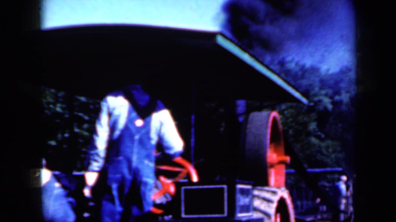 A man in blue overalls stands close to a red flywheel as smoke rises from the machine in this 1959 Iowa home-movie frame.