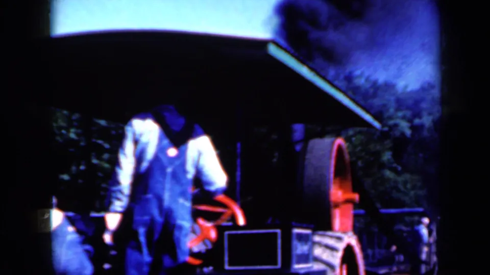 A man in blue overalls stands close to a red flywheel as smoke rises from the machine in this 1959 Iowa home-movie frame.