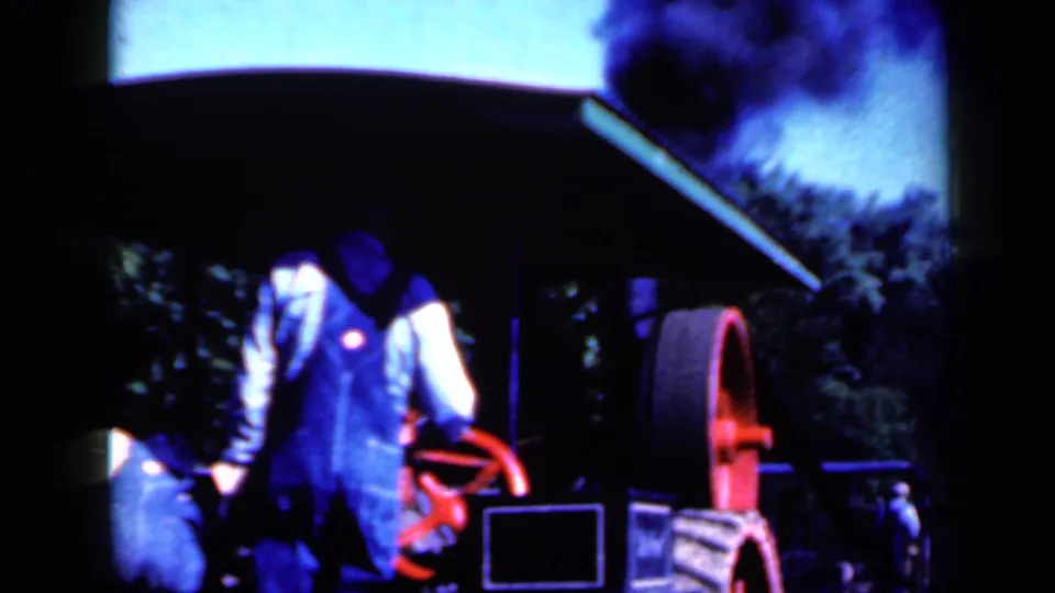 Workers gather around the steam-powered machine, with one man near the red wheel and smoke visible against the sky.