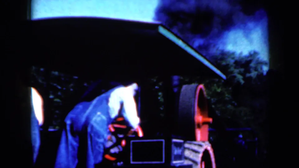 The operator bends toward the machine near the red wheel as smoke rises, with another worker partly visible at the edge of the frame.