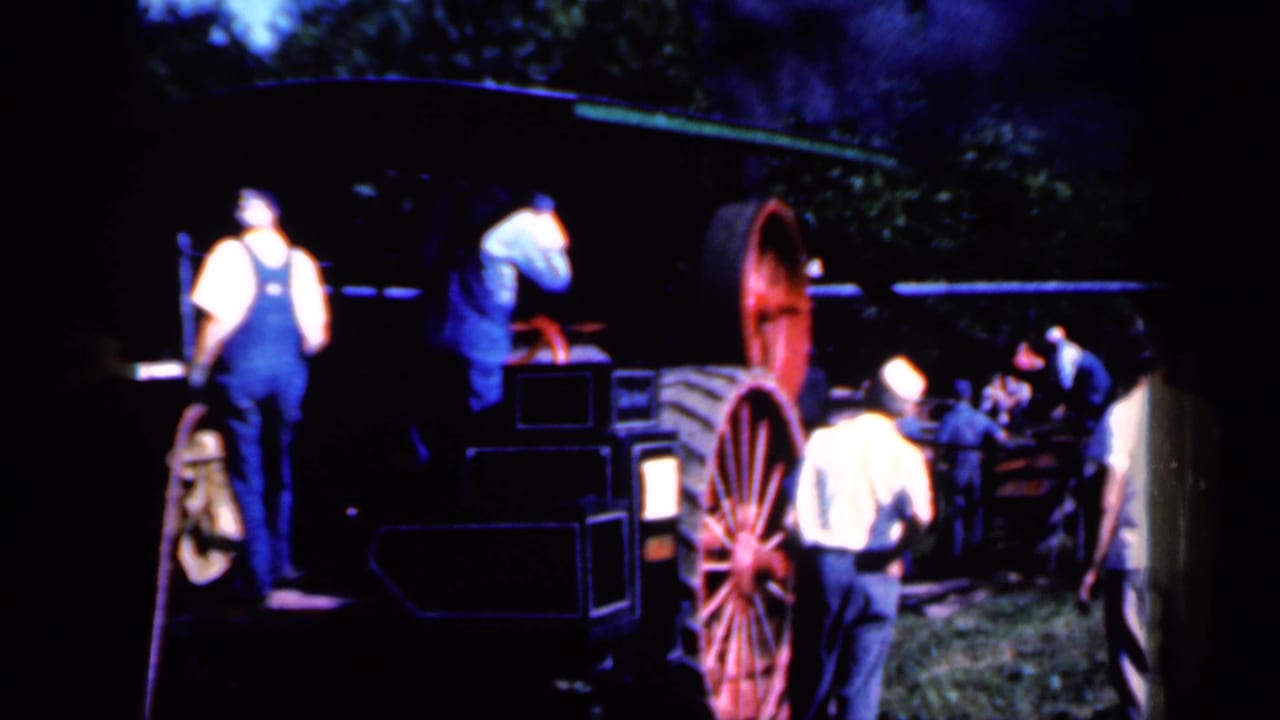 Men in work clothes stand beside a large farm machine with red spoked wheels outdoors.