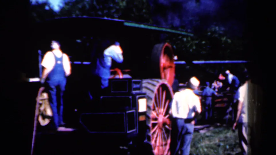 In Iowa in 1959, men in overalls and work shirts gather near a large tractor or farm machine with red spoked wheels under dim outdoor light.