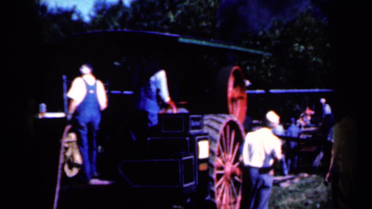 A 1959 Iowa home-movie frame shows men around a dark tractor-like machine with a large red spoked wheel and trees behind them.