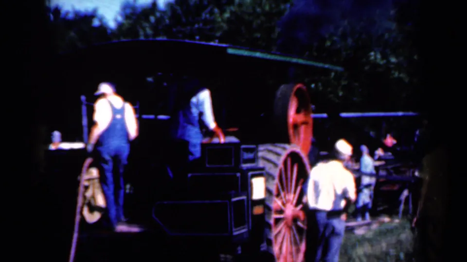 Men in mid-century work clothing appear around farm equipment with red spoked wheels in an Iowa rural setting, filmed on 8mm in 1959.