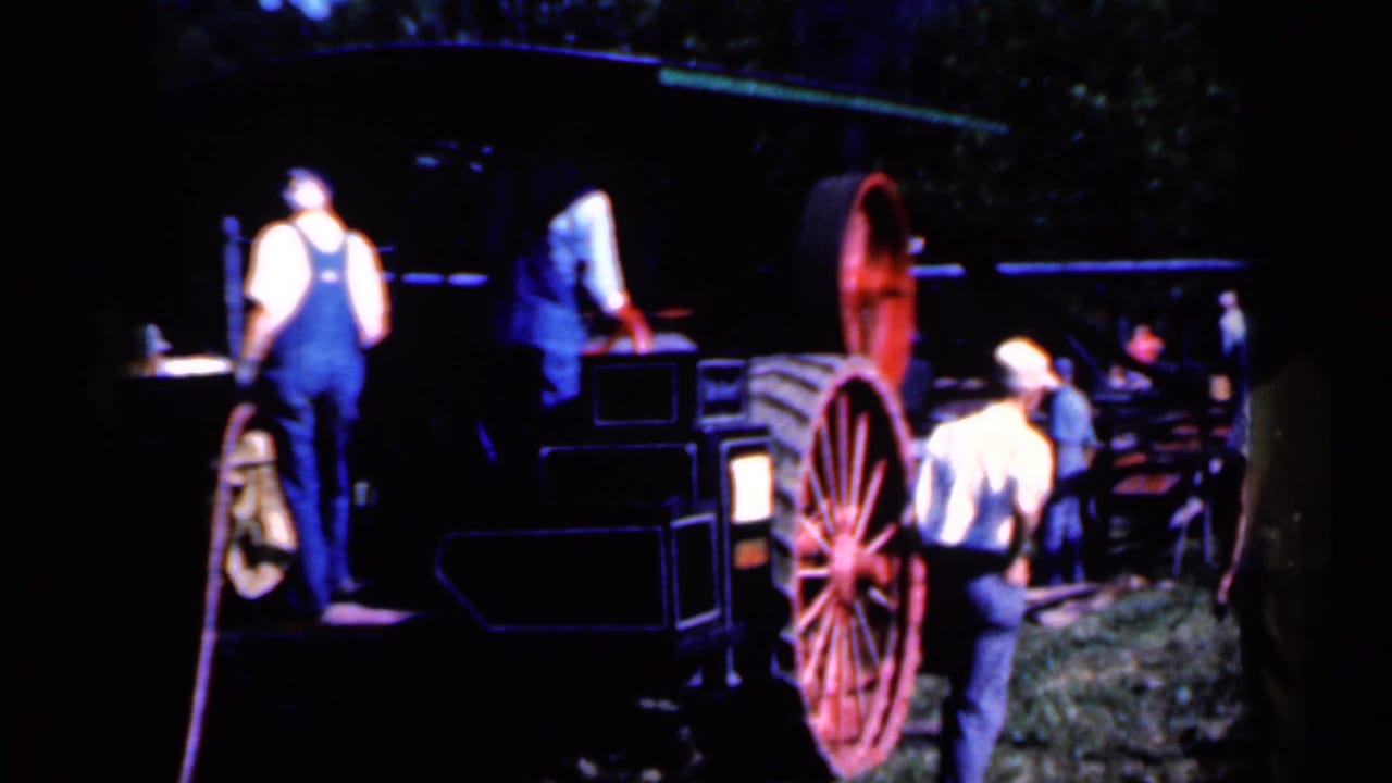 Men in overalls stand beside outdoor sawmill machinery and a large red spoked wheel.
