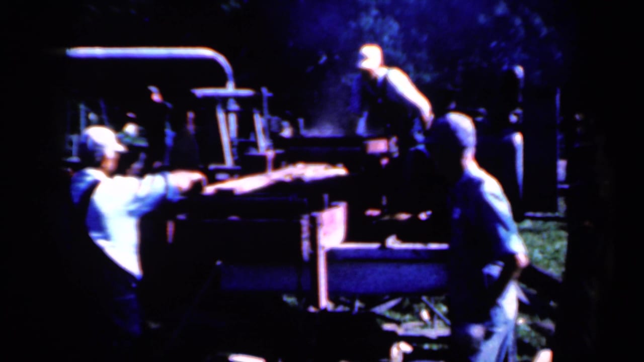 1959 Iowa rural work frame with men guiding timber into or across a large outdoor sawmill machine.