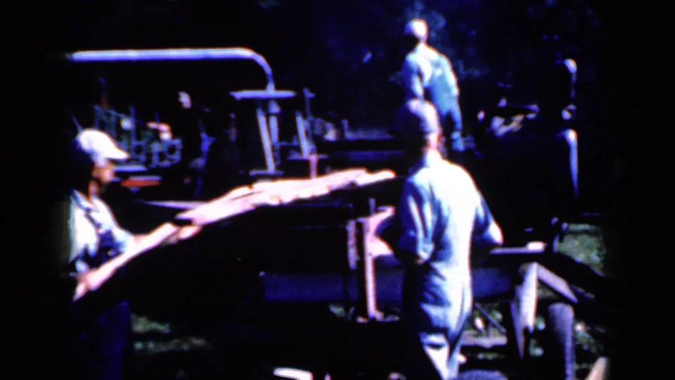 1959 Iowa 8mm frame of workers in caps and overalls guiding a log near rural sawmill equipment.