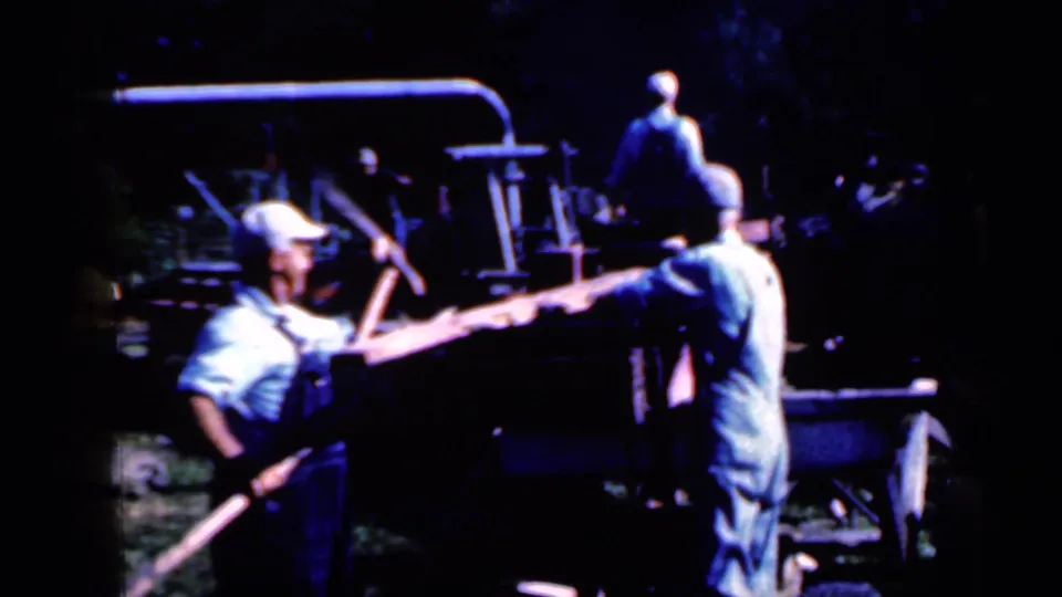 1959 Iowa home-movie frame showing men handling timber at an outdoor sawmill under high-contrast exposure.