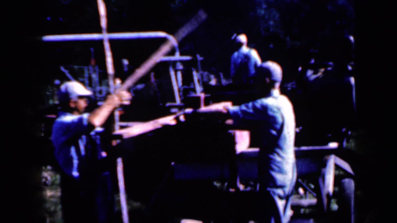 Men handle long wooden posts beside outdoor machinery in deep shade.