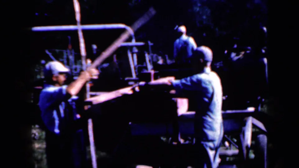 In Iowa 1959 home-movie footage, men in caps appear to guide wooden posts near a fence-post processing machine.