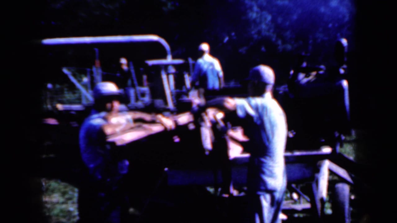 Men in work clothes appear to guide wooden posts into or along a rural processing machine in Iowa, 1959.