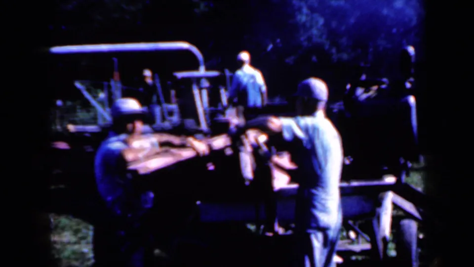 Men in work clothes appear to guide wooden posts into or along a rural processing machine in Iowa, 1959.