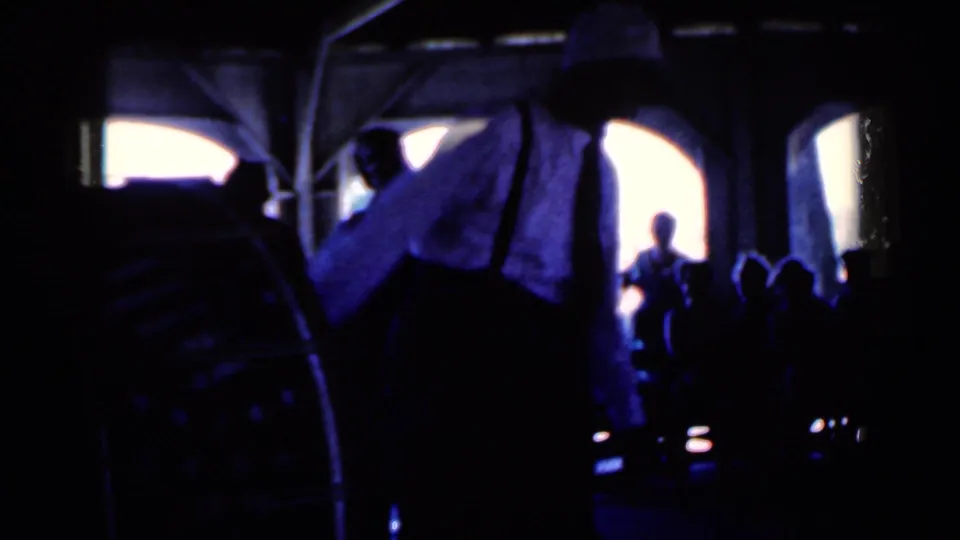 1959 Iowa home-movie frame showing a dim party room with a man in suspenders, seated silhouettes, and bright arched openings.